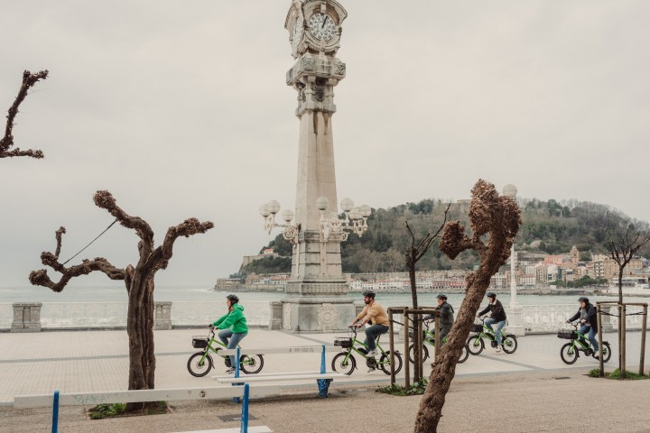 Four people riding bicycles along a seaside promenade with trees and a clock tower.